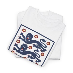 Puede incluir: Camiseta blanca con un diseño azul marino y rojo. El diseño incluye dos leones azul marino y detalles florales rojos dentro de un cuadrado con borde azul marino. La camiseta está doblada y apilada sobre otra camiseta blanca.