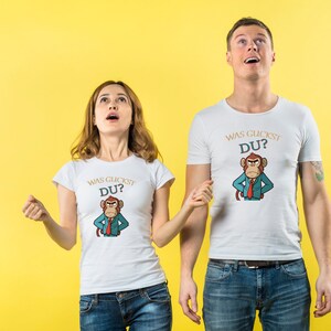 May include: Two people wearing white t-shirts with a cartoon monkey design and the text "WAS GUCKST DU?" in gold lettering. The monkey is wearing a suit and tie. The background is a solid yellow.