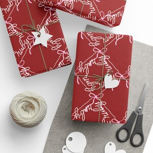 May include: Three wrapped gifts with red paper featuring a white and pink script design, tied with twine and white paper tags. A ball of twine, scissors, and blank tags are also visible on a white and gray surface.