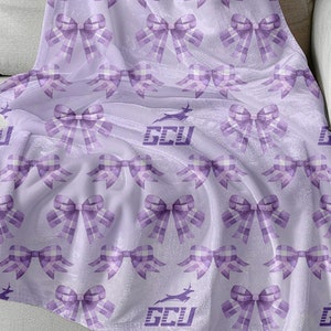 以下が含まれることがあります： 紫色のフリース素材の柔らかい毛布で、紫色のリボンと「GCU」という文字が紫色のフォントで繰り返しパターンになっています。毛布は薄い灰色のソファにかけられています。