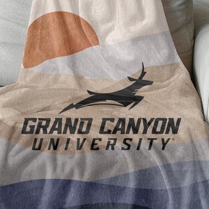 以下が含まれることがあります： 夕焼けと山岳風景のデザインが施された、柔らかなベージュの毛布。毛布には、跳ね上がるオリックスのシルエットが描かれたグランドキャニオン大学のロゴがデザインされています。