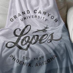 以下が含まれることがあります： グランドキャニオン大学のロゴが入った白と青のグラデーションの毛布。ロゴは、スタイリッシュなフォントで「Lopes」という単語を囲む円の中に「Grand Canyon University」という単語が書かれています。「Est. 1949 Phoenix, Arizona」というテキストがロゴの下にあります。