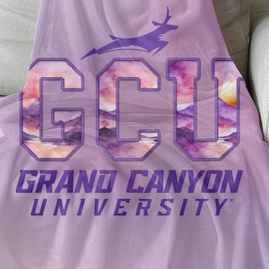 以下が含まれることがあります： グレーのソファにかけられた、ライラックと紫色のブランケット。「GCU GRAND CANYON UNIVERSITY」の文字が紫で、文字の中に風景デザインと紫色のアンテロープロゴが描かれています。