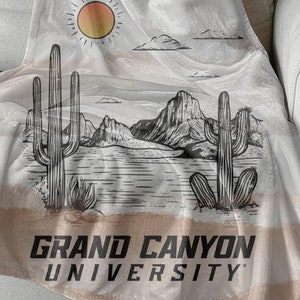 以下が含まれることがあります： 砂漠の風景のデザインが施された、タンとブラウンのフリースの毛布。毛布には、太陽、山、サボテン、そして「グランドキャニオン大学」の文字が描かれています。
