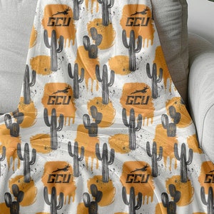 以下が含まれることがあります： 黒いサボテンとオレンジ色のペイントの飛沫が繰り返されるパターンが施された白い毛布。パターン全体に「GCU」という文字が繰り返されています。