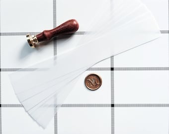 May include: A set of white wax seal paper sheets with a wooden handle wax seal stamp and a bronze wax seal with the letter "M" on a white and black checkered surface.