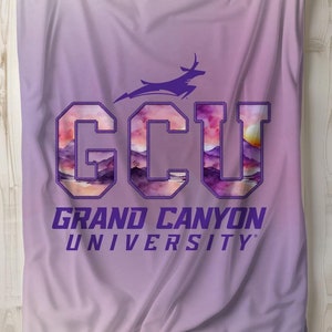 以下が含まれることがあります： 紫とピンクの水彩画のデザインで、紫色の輪郭で「GCU」の文字が描かれています。文字は、山と夕日の水彩画のデザインで満たされています。文字の下には「グランドキャニオン大学」の文字があります。