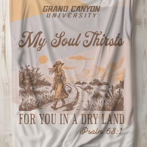 以下が含まれることがあります： 砂漠の風景の中で、土の道を歩く女性を描いた、茶色とオレンジのデザインが施された白い毛布。毛布には「My Soul Thirsts」と「For You in a Dry Land」の文字が印刷されており、さらに「Grand Canyon University」と「Psalm 63:1」の文字も印刷されています。