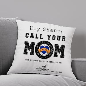 May include: White cushion with the message "Hey Shane, CALL YOUR MOM" in black text. The word "MOM" is in large, distressed black letters with a blue heart tattoo design in the center. The cushion is endorsed by Grand Canyon University.