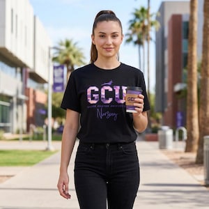 Op de afbeelding: Een zwart T-shirt met korte mouwen met de paarse en witte tekst GCU Grand Canyon University Nursing en een kleine antilope-afbeelding boven de letters. Het shirt wordt gedragen met een zwarte spijkerbroek en een koffiebeker met paarse hoes.