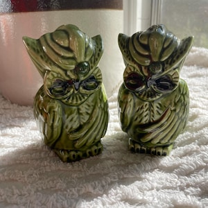 May include: A pair of green ceramic owl salt and pepper shakers. The owls have black eyes and are sitting on a white fluffy surface.