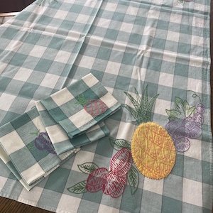 May include: A light blue and white checkered tablecloth and matching napkins. The tablecloth and napkins are decorated with embroidered fruits, including pineapples, grapes, and other fruits. The tablecloth is folded on a wooden surface.