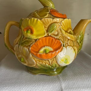 May include: A ceramic teapot with a floral design. The teapot is decorated with yellow, orange, and white flowers. The teapot has a green lid and a green handle.