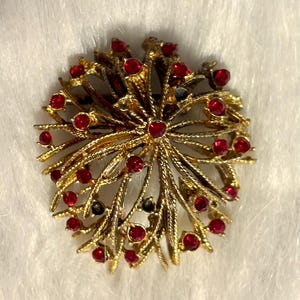 May include: A gold-toned brooch with a radial design, featuring intricate branches and red gemstone accents. The brooch has a textured finish and a central cluster of red stones, with additional stones along the branches.