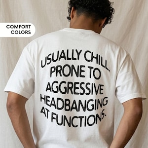 May include: White t-shirt with the text "USUALLY CHILL PRONE TO AGGRESSIVE HEADBANGING AT FUNCTIONS" in black. The shirt is paired with black jeans. The words "COMFORT COLORS" are in a white box in the upper left corner.
