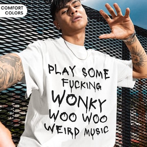 May include: White t-shirt with the text "PLAY SOME FUCKING WONKY WOO WOO WEIRD MUSIC" in black. The shirt is part of the "COMFORT COLORS" collection. The person is wearing black shorts and a silver chain.