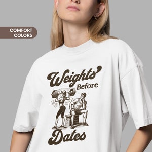 May include: White t-shirt with a brown graphic of a man and woman lifting weights. The text "Weights Before Dates" is printed on the shirt.