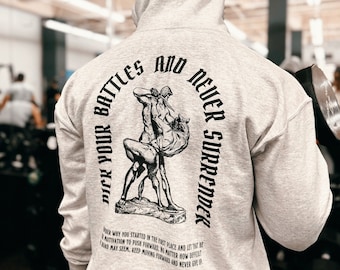 Puede incluir: Una sudadera con capucha gris claro que presenta un gráfico negro en la parte posterior. El diseño muestra a una figura luchando contra un toro con el texto "PICK YOUR BATTLES AND NEVER SURRENDER" y un párrafo motivacional debajo.