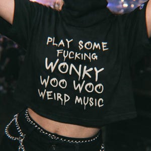 K&ouml;nnte beinhalten: Schwarzes Crop-Top mit dem wei&szlig;en Text "PLAY SOME FUCKING WONKY WOO WOO WEIRD MUSIC". Das Outfit umfasst schwarze Shorts und einen silbernen Ketten-G&uuml;rtel.