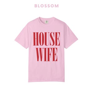 May include: A light pink t-shirt with the word "HOUSEWIFE" printed in bold red letters. The word "BLOSSOM" is printed in a lighter pink above the shirt.