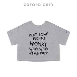 K&ouml;nnte beinhalten: Oxfordgraues Crop-Top mit dem Text "PLAY SOME FUCKING WONKY WOO WOO WEIRD MUSIC" in Schwarz. Das Shirt hat kurze &Auml;rmel und einen Rundhalsausschnitt. Ein Champion-Logo ist auf dem linken &Auml;rmel zu sehen.