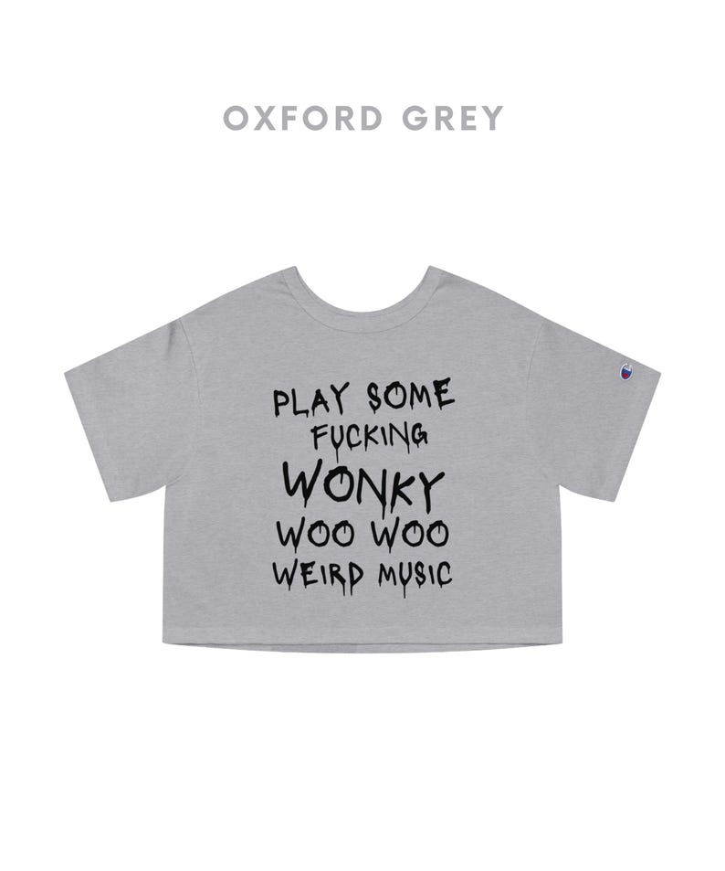 K&ouml;nnte beinhalten: Oxfordgraues Crop-Top mit dem Text "PLAY SOME FUCKING WONKY WOO WOO WEIRD MUSIC" in Schwarz. Das Shirt hat kurze &Auml;rmel und einen Rundhalsausschnitt. Ein Champion-Logo ist auf dem linken &Auml;rmel zu sehen.