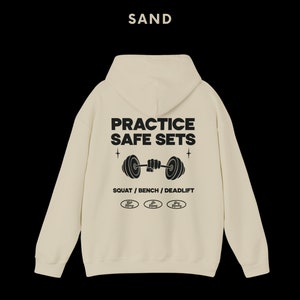 K&ouml;nnte beinhalten: Ein sandfarbener Kapuzenpullover mit dem Text "PRACTICE SAFE SETS" und einer Grafik einer Person, die eine Langhantel hebt. Der Text "SQUAT / BENCH / DEADLIFT" befindet sich unter der Grafik. Drei kleine Kreise mit dem Text "Safe Lifting" befinden sich unter dem Text.
