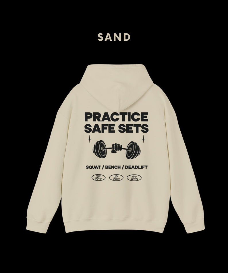 K&ouml;nnte beinhalten: Ein sandfarbener Kapuzenpullover mit dem Text "PRACTICE SAFE SETS" und einer Grafik einer Person, die eine Langhantel hebt. Der Text "SQUAT / BENCH / DEADLIFT" befindet sich unter der Grafik. Drei kleine Kreise mit dem Text "Safe Lifting" befinden sich unter dem Text.