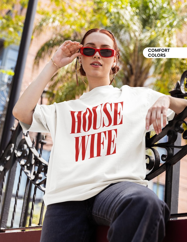 May include: A white t-shirt with the red text "HOUSE WIFE". The person in the image is wearing red sunglasses and dark jeans. The words "COMFORT COLORS" are in a white box in the upper right corner.