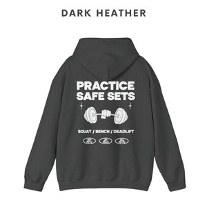 K&ouml;nnte beinhalten: Dunkelgrauer Kapuzenpullover mit einem wei&szlig;en Grafikdruck, der "Practice Safe Sets" mit einer Langhantel und den Worten "Squat / Bench / Deadlift" darunter sagt. Die Grafik enth&auml;lt auch drei kleine Kreise mit den Worten "Safe Heavy" darin.