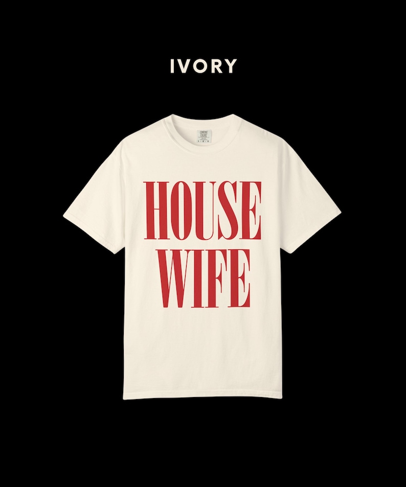 May include: Ivory-colored t-shirt with the word "HOUSEWIFE" printed in red. The text is vertically stacked and centered on the shirt. The shirt has a crew neck and short sleeves. The background is black.