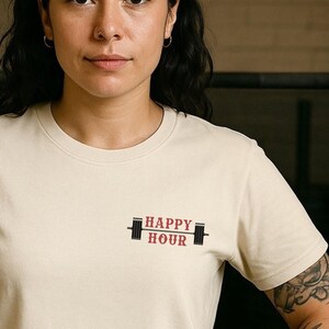 Op de afbeelding: Cr&egrave;mekleurig T-shirt met de tekst "HAPPY HOUR" in rood en zwart, vergezeld van een haltergrafiek. Het shirt wordt gedragen door een persoon met donker haar en een neusring.