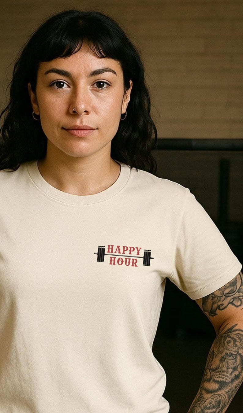 Op de afbeelding: Cr&egrave;mekleurig T-shirt met de tekst "HAPPY HOUR" in rood en zwart, vergezeld van een haltergrafiek. Het shirt wordt gedragen door een persoon met donker haar en een neusring.