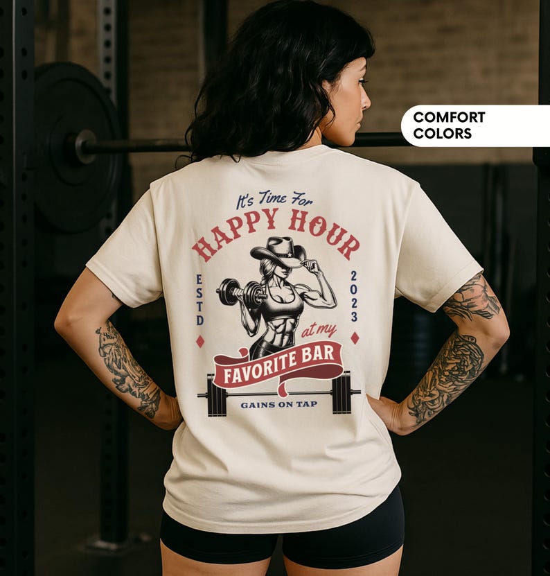 Op de afbeelding: Cr&egrave;mekleurig T-shirt met een afbeelding van een gewichtheffende cowgirl en de tekst "It's Time For Happy Hour at my Favorite Bar". Het ontwerp bevat het jaar 2023 en de zin "Gains on Tap".