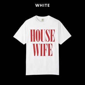 May include: White t-shirt with the word "HOUSEWIFE" printed in red. The text is vertically aligned and centered on the shirt. The word "WHITE" is printed above the shirt. The shirt has short sleeves and a crew neck.