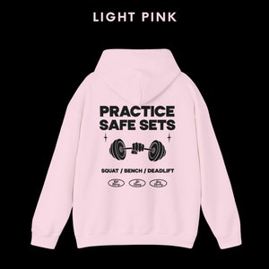 K&ouml;nnte beinhalten: Hellrosa Kapuzenpullover mit dem Text "PRACTICE SAFE SETS" und einer Grafik einer Person, die eine Langhantel hebt. Der Text "SQUAT / BENCH / DEADLIFT" befindet sich unter der Grafik. Drei kleine Kreise mit dem Text "Safe Lifting" befinden sich unter dem Text.
