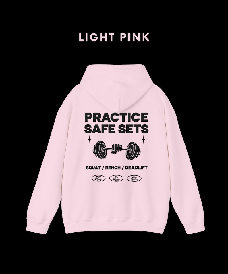 K&ouml;nnte beinhalten: Hellrosa Kapuzenpullover mit dem Text "PRACTICE SAFE SETS" und einer Grafik einer Person, die eine Langhantel hebt. Der Text "SQUAT / BENCH / DEADLIFT" befindet sich unter der Grafik. Drei kleine Kreise mit dem Text "Safe Lifting" befinden sich unter dem Text.
