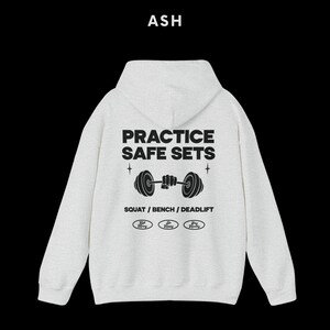K&ouml;nnte beinhalten: Aschgrauer Kapuzenpullover mit dem Text "Practice Safe Sets" und einer Grafik einer Person, die eine Langhantel hebt. Der Text "Squat / Bench / Deadlift" befindet sich unter der Grafik. Drei kleine Kreise mit dem Text "Stay Strong" befinden sich unter dem Text.