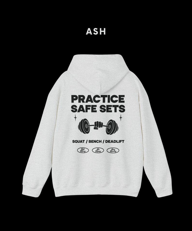 K&ouml;nnte beinhalten: Aschgrauer Kapuzenpullover mit dem Text "Practice Safe Sets" und einer Grafik einer Person, die eine Langhantel hebt. Der Text "Squat / Bench / Deadlift" befindet sich unter der Grafik. Drei kleine Kreise mit dem Text "Stay Strong" befinden sich unter dem Text.