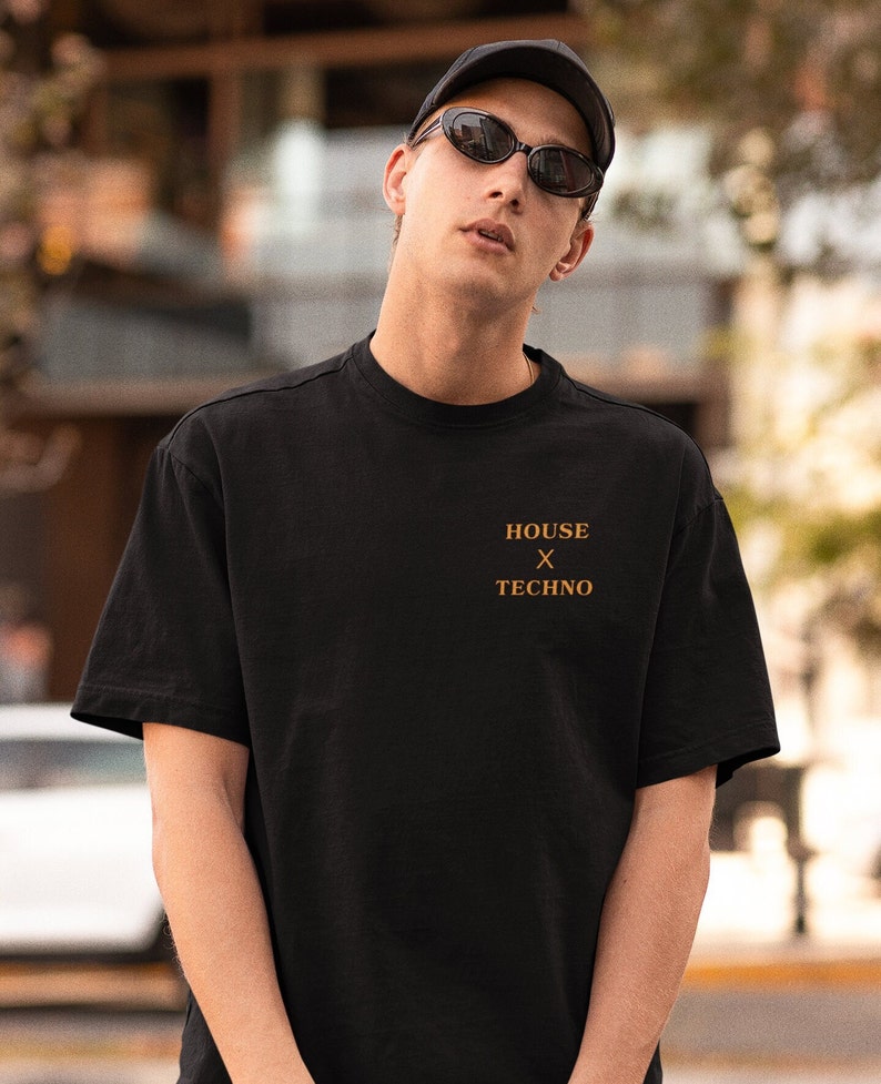 Pu&ograve; includere: Una persona indossa una maglietta nera con la scritta "HOUSE x TECHNO" stampata sul davanti. La persona indossa un berretto nero e occhiali da sole.