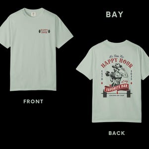 Op de afbeelding: Lichtgroen t-shirt met "HAPPY HOUR" en halterlogo op de voorkant. De achterkant heeft een afbeelding van een vrouw die gewichten heft, de tekst "It's Time For HAPPY HOUR at my FAVORITE BAR" en "GAINS ON TAP".
