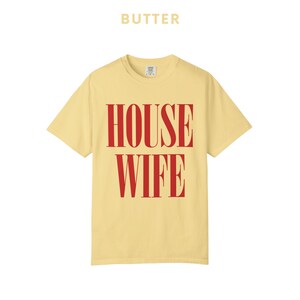 May include: Butter-colored t-shirt with the word "BUTTER" printed above the large red text "HOUSE WIFE". The shirt has a crew neck and short sleeves. The shirt is a solid color.