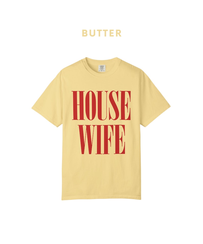 May include: Butter-colored t-shirt with the word "BUTTER" printed above the large red text "HOUSE WIFE". The shirt has a crew neck and short sleeves. The shirt is a solid color.