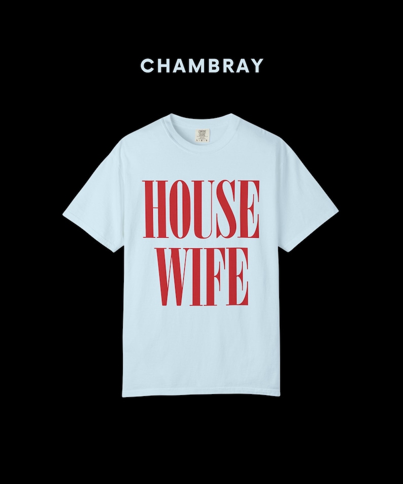 May include: Light blue t-shirt with the word "HOUSEWIFE" in large, red, bold letters. The word "CHAMBRAY" is above the shirt. The shirt has short sleeves and a crew neck.