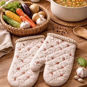 May include: Cream-coloured quilted oven mitts with a strawberry pattern, next to a basket of fresh vegetables and a pot of soup. The mitts have a diamond pattern and small red strawberry designs.