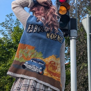 May include: A woman wearing a blue and white crocheted sweater with a car and the text "Everybody Does It" on the back. She is also wearing a plaid skirt.