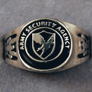May include: A silver ring with a black enamel inlay featuring the words "Army Security Agency" and a stylized bird inside a shield.