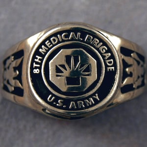 Może przedstawiać: Srebrny pierścień z czarną emalią. Pierścień zawiera napis "8th Medical Brigade U.S. Army" i symbol medyczny.