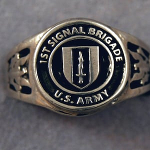 May include: A silver ring with a black enamel center featuring the inscription "1st Signal Brigade U.S. Army". The center design includes a shield with a sword.