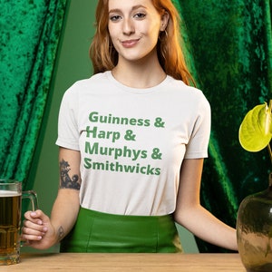May include: A woman wearing a white t-shirt with green text that reads "Guinness & Harp & Murphys & Smithwicks" and a green skirt. She is holding a glass of beer in her right hand. The background is green.